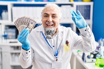 Middle age man with grey hair working at scientist laboratory holding money doing ok sign with fingers, smiling friendly gesturing excellent symbol