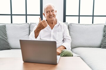 Senior man using laptop at home sitting on the sofa smiling with happy face looking and pointing to the side with thumb up.