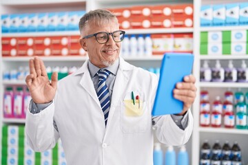Hispanic man with grey hair working at pharmacy drugstore doing video call with tablet looking positive and happy standing and smiling with a confident smile showing teeth