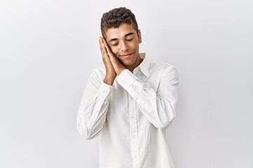 Young handsome hispanic man standing over isolated background sleeping tired dreaming and posing with hands together while smiling with closed eyes.