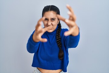 Young hispanic woman standing over isolated background shouting frustrated with rage, hands trying to strangle, yelling mad