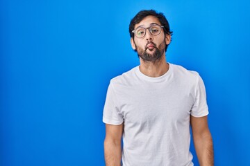 Handsome latin man standing over blue background making fish face with lips, crazy and comical gesture. funny expression.