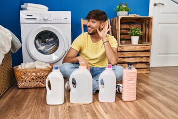 Arab man with beard doing laundry sitting on the floor with detergent bottle smiling with hand over ear listening and hearing to rumor or gossip. deafness concept.