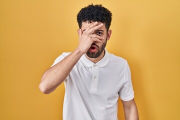 Arab man standing over yellow background peeking in shock covering face and eyes with hand, looking...