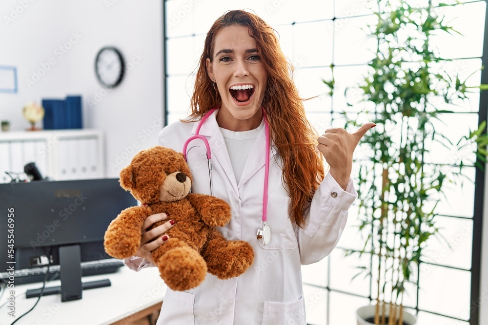 Poster young redhead pedriatician woman holding teddy bear pointing thumb up to the side smiling happy with