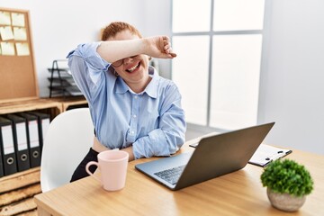 Young redhead woman working at the office using computer laptop covering eyes with arm smiling cheerful and funny. blind concept.