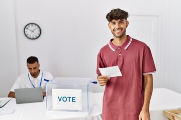 Young hispanic man voting putting envelop in ballot box looking positive and happy standing and smiling with a confident smile showing teeth