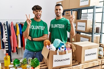 Young gay couple wearing volunteer t shirt at donations stand smiling with happy face winking at...