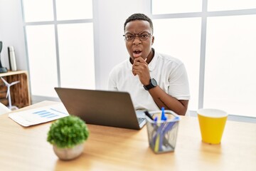 Young african man working at the office using computer laptop looking fascinated with disbelief, surprise and amazed expression with hands on chin