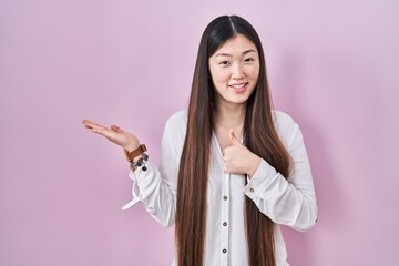 Chinese young woman standing over pink background showing palm hand and doing ok gesture with...