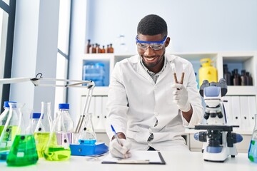 Young african american man wearing scientist uniform writing on clipboard holding test tubes at laboratory