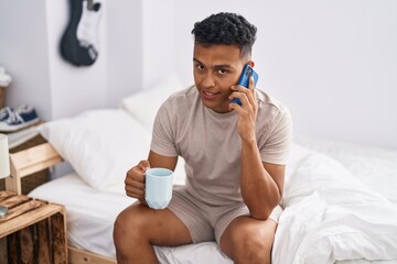 Obraz na płótnie Canvas Young latin man talking on smartphone drinking coffee at bedroom
