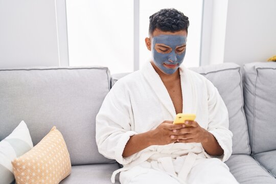 Young latin man relaxed on sofa with facial mask treatment using smartphone at home