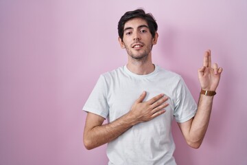 Young hispanic man standing over pink background smiling swearing with hand on chest and fingers up, making a loyalty promise oath