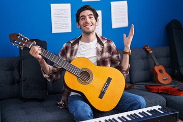 Young hispanic man playing classic guitar at music studio celebrating victory with happy smile and winner expression with raised hands