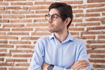 Young hispanic man standing over brick wall background looking to the side with arms crossed convinced and confident