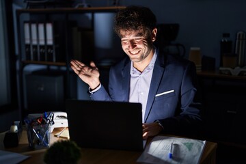 Hispanic young man working at the office at night smiling cheerful presenting and pointing with palm of hand looking at the camera.