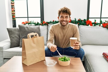 Young hispanic man eating take away salad sitting by christmas decor at home