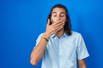 Young hispanic man standing over blue background bored yawning tired covering mouth with hand. restless and sleepiness.