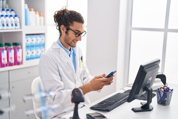Young hispanic man pharmacist using smartphone working at pharmacy