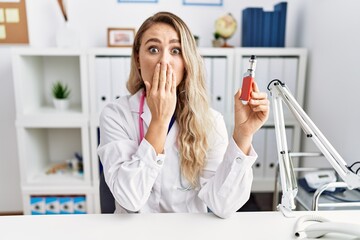 Young beautiful doctor woman holding electronic cigarette at the clinic covering mouth with hand,...