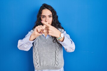 Young brunette woman standing over blue background rejection expression crossing fingers doing...