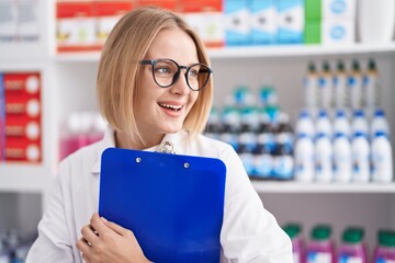 Young blonde woman pharmacist smiling confident holding clipboard at pharmacy