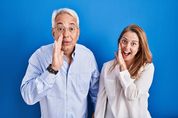 Middle age hispanic couple standing over blue background hand on mouth telling secret rumor,...