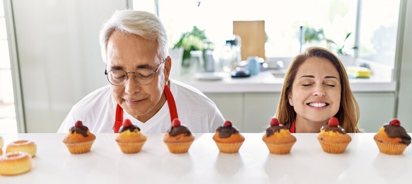 Middle age hispanic couple smiling happy smelling muffins at the kitchen.