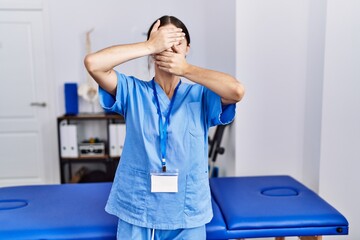 Obraz premium Young hispanic woman wearing physiotherapist uniform standing at clinic covering eyes and mouth with hands, surprised and shocked. hiding emotion