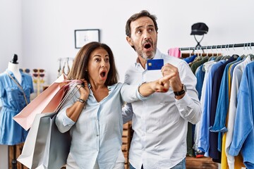Hispanic middle age couple holding shopping bags and credit card pointing with finger surprised ahead, open mouth amazed expression, something on the front