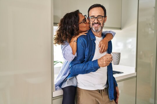 Middle age hispanic couple kissing and hugging each other drinking coffee at kitchen