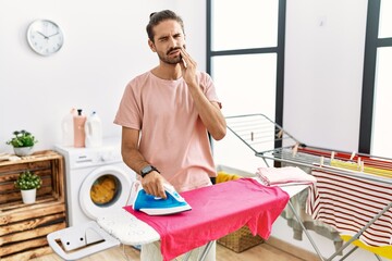 Young hispanic man ironing clothes at home touching mouth with hand with painful expression because of toothache or dental illness on teeth. dentist concept.