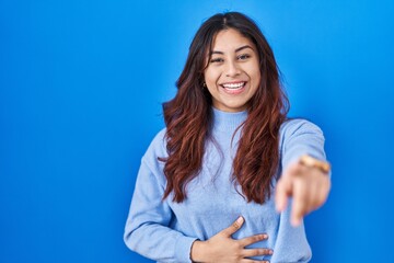 Hispanic young woman standing over blue background laughing at you, pointing finger to the camera with hand over body, shame expression