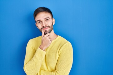 Hispanic man standing over blue background looking confident at the camera smiling with crossed arms and hand raised on chin. thinking positive.