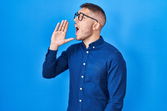 Young hispanic man wearing glasses over blue background shouting and screaming loud to side with hand on mouth. communication concept.
