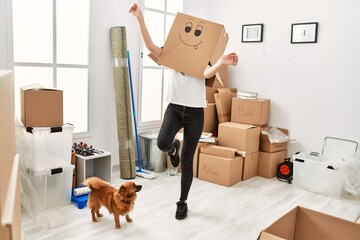 Young hispanic woman wearing funny cardboard on head playing with dog at new home