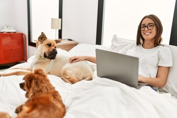 Obraz premium Young hispanic woman using laptop sitting on bed with dogs at bedroom