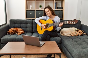 Young hispanic woman having online guitar class sitting on sofa with dogs at home