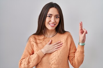 Young brunette woman standing over white background smiling swearing with hand on chest and fingers up, making a loyalty promise oath