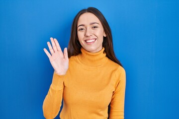 Young brunette woman standing over blue background waiving saying hello happy and smiling, friendly welcome gesture © Krakenimages.com