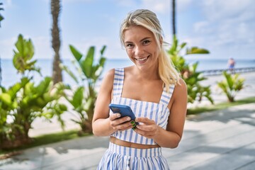 Young blonde girl smiling happy using smartphone at the promenade.