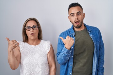 Hispanic mother and son standing together surprised pointing with hand finger to the side, open mouth amazed expression.