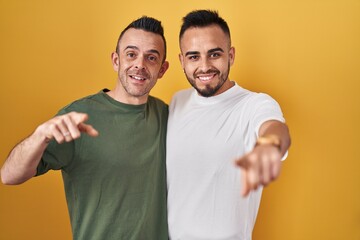 Homosexual couple standing over yellow background pointing to you and the camera with fingers, smiling positive and cheerful