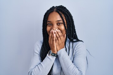 African american woman standing over blue background laughing and embarrassed giggle covering mouth...
