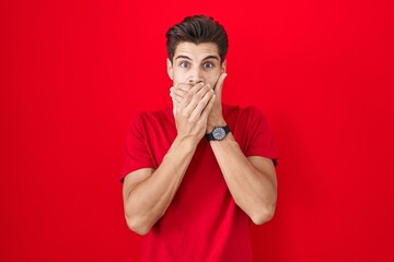 Young hispanic man standing over red background shocked covering mouth with hands for mistake....