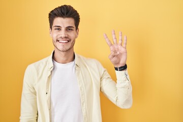 Young hispanic man standing over yellow background showing and pointing up with fingers number four...