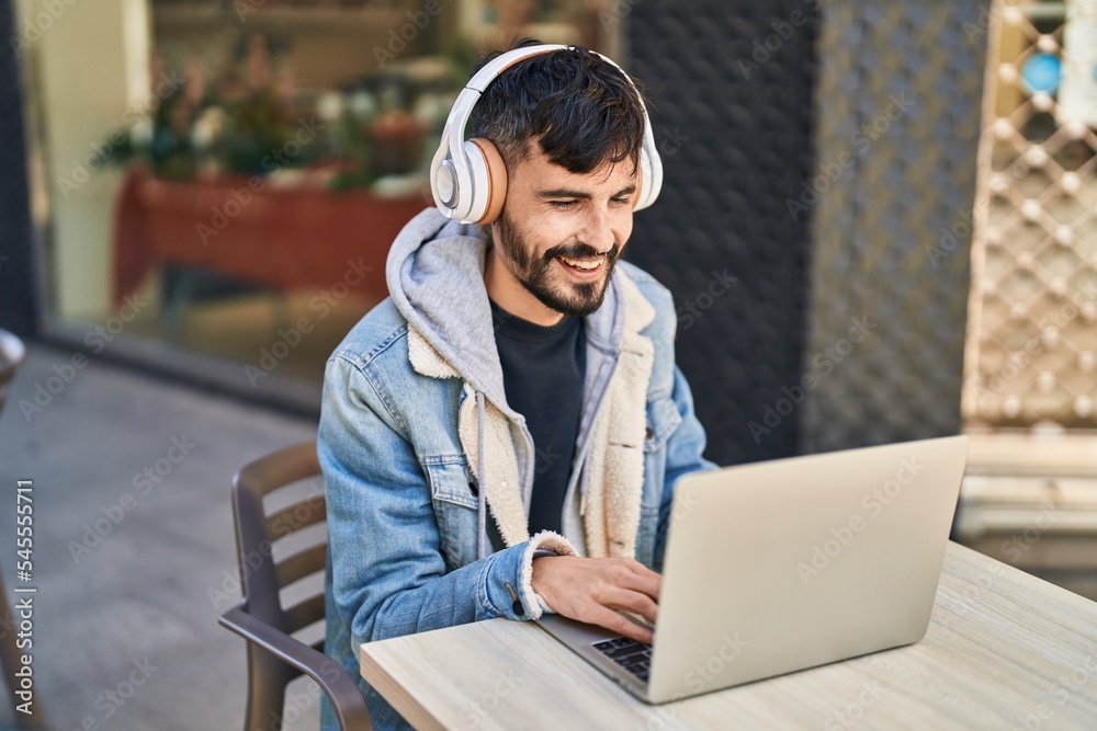 Poster young hispanic man using laptop and headphones sitting on table at coffee shop terrace