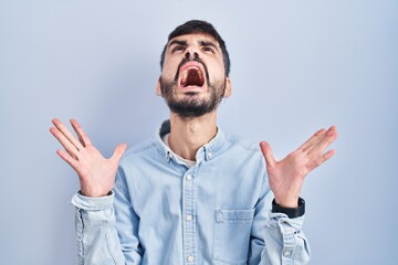 Young hispanic man with beard standing over blue background crazy and mad shouting and yelling with aggressive expression and arms raised. frustration concept.