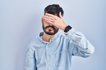 Young hispanic man with beard standing over blue background covering eyes with hand, looking serious and sad. sightless, hiding and rejection concept